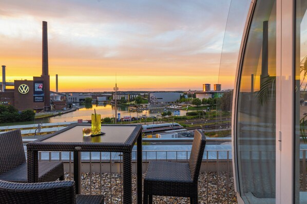 Une terrasse dotée de meubles en osier, d’une table et offrant une vue sur une rivière, des bâtiments et un pont, le tout baigné par les couleurs du coucher de soleil.