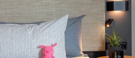 A bed with patterned pillows, a small round table with a plant and a mug, and a wall-mounted light fixture.