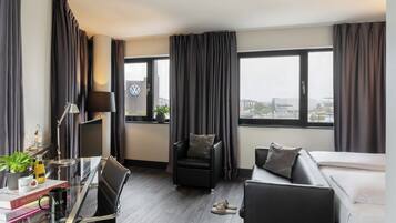 A modern hotel room with a glass desk, a black leather chair, a sofa, and a bed. There is a large window with curtains, a lamp, and a view of a cityscape.