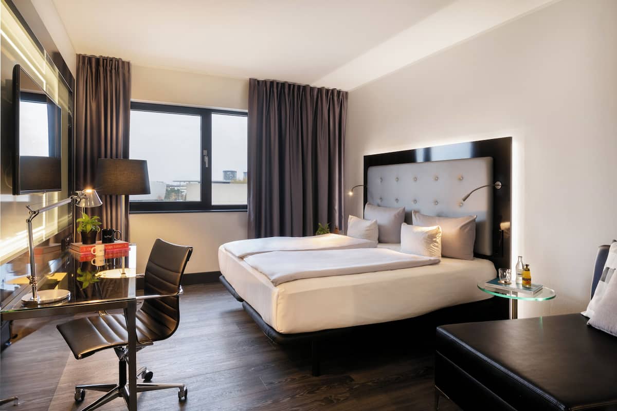 A modern hotel room with a large bed, a desk with a chair, a glass table, and a view of the city.