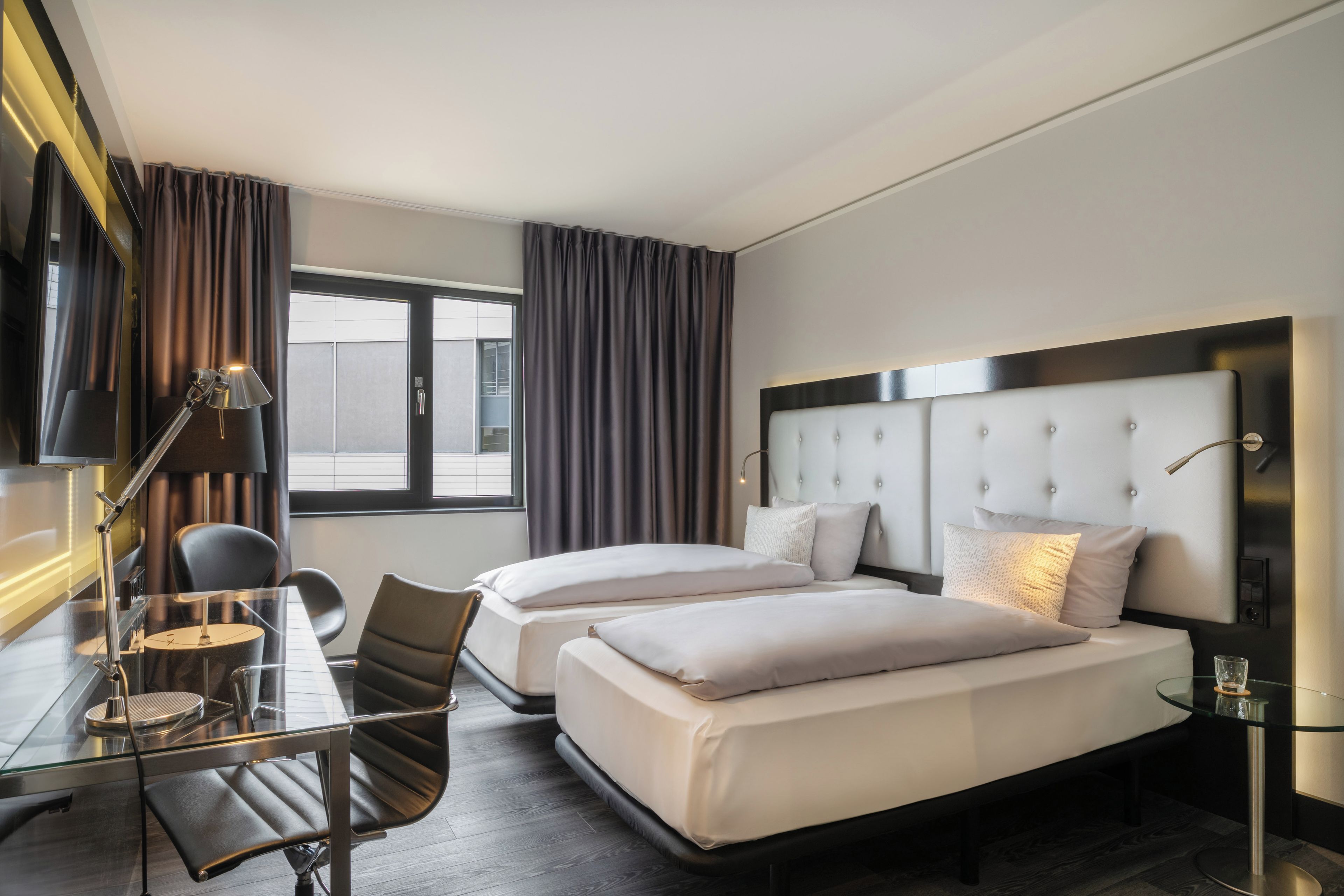 Quarto de hotel moderno com duas camas, uma escrivaninha com luminária, uma cadeira e uma TV.