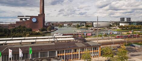 A large industrial building with a tall chimney, a river with boats, and a modern train station with tracks and a red train.