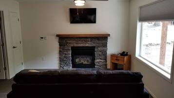 A living room with a stone fireplace, a television mounted on the wall, and a window with a view of the outdoors.