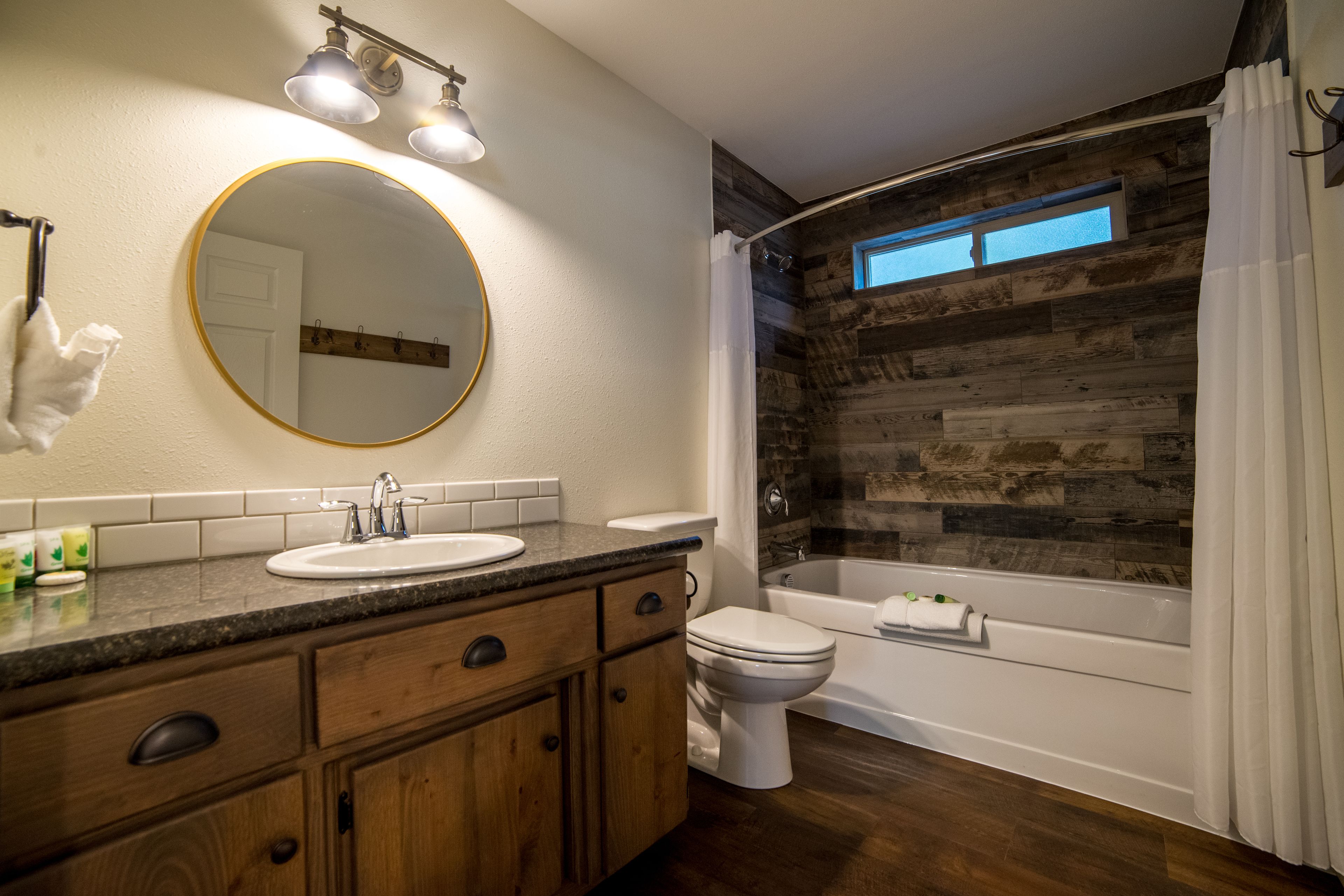 A bathroom with a wooden wall, a round mirror, a sink, a toilet, and a bathtub.