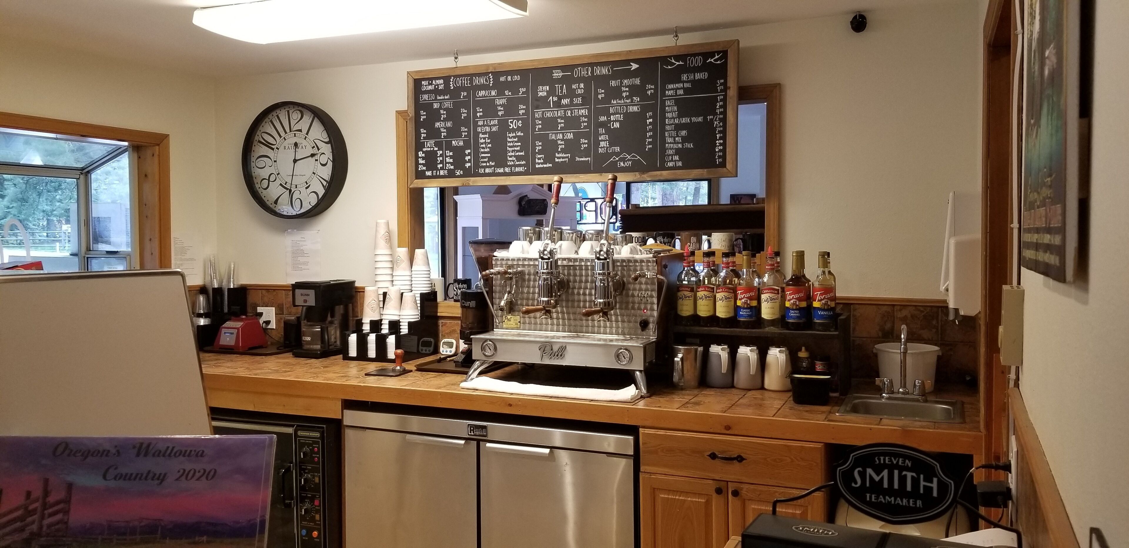 Interior de una cafetería con una máquina de café, una pizarra de menú y varias tazas y botellas de café.