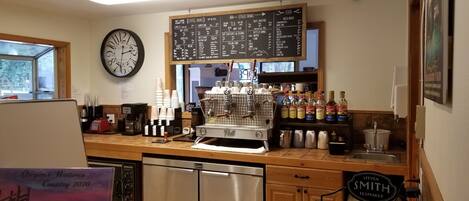 Interior de una cafetería con una máquina de café, una pizarra de menú y varias tazas y botellas de café.