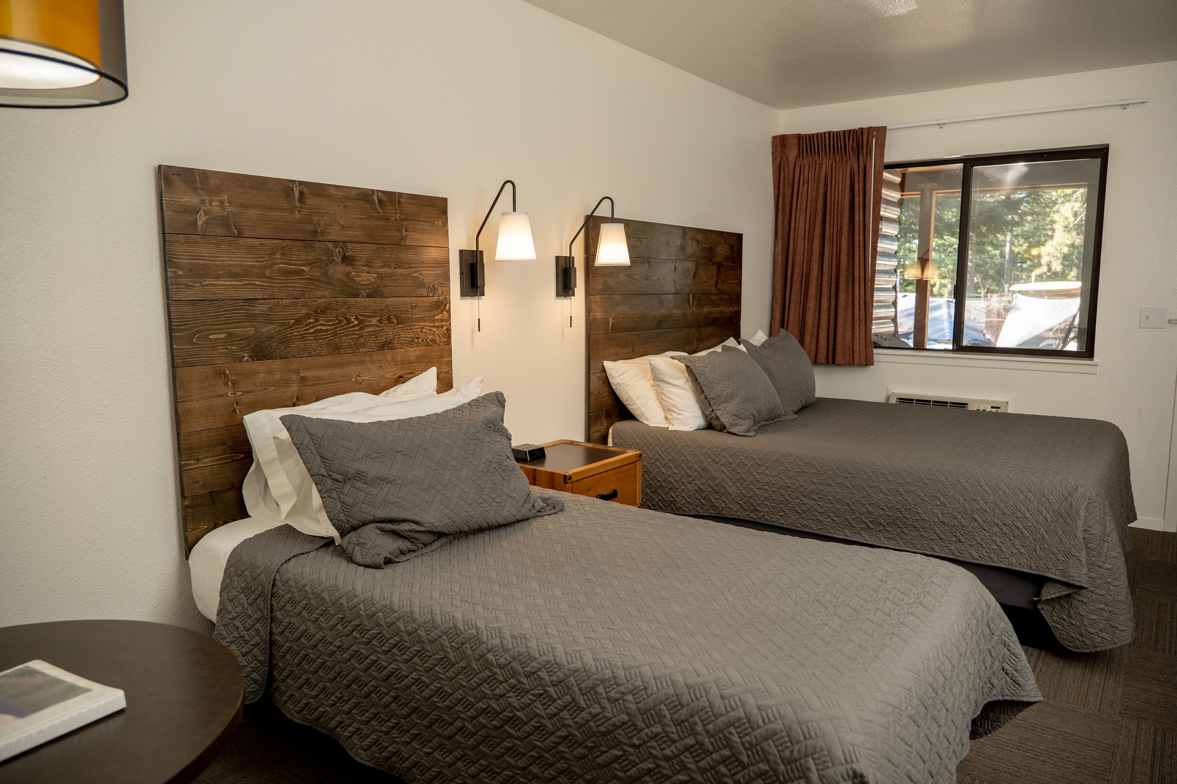 Une chambre d’hôtel avec deux lits, des têtes de lit en bois, des lampes de chevet, une fenêtre avec des rideaux et une petite table sur laquelle se trouve un livre.