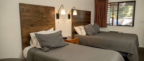 Une chambre d’hôtel avec deux lits, des têtes de lit en bois, des lampes de chevet, une fenêtre avec des rideaux et une petite table sur laquelle se trouve un livre.