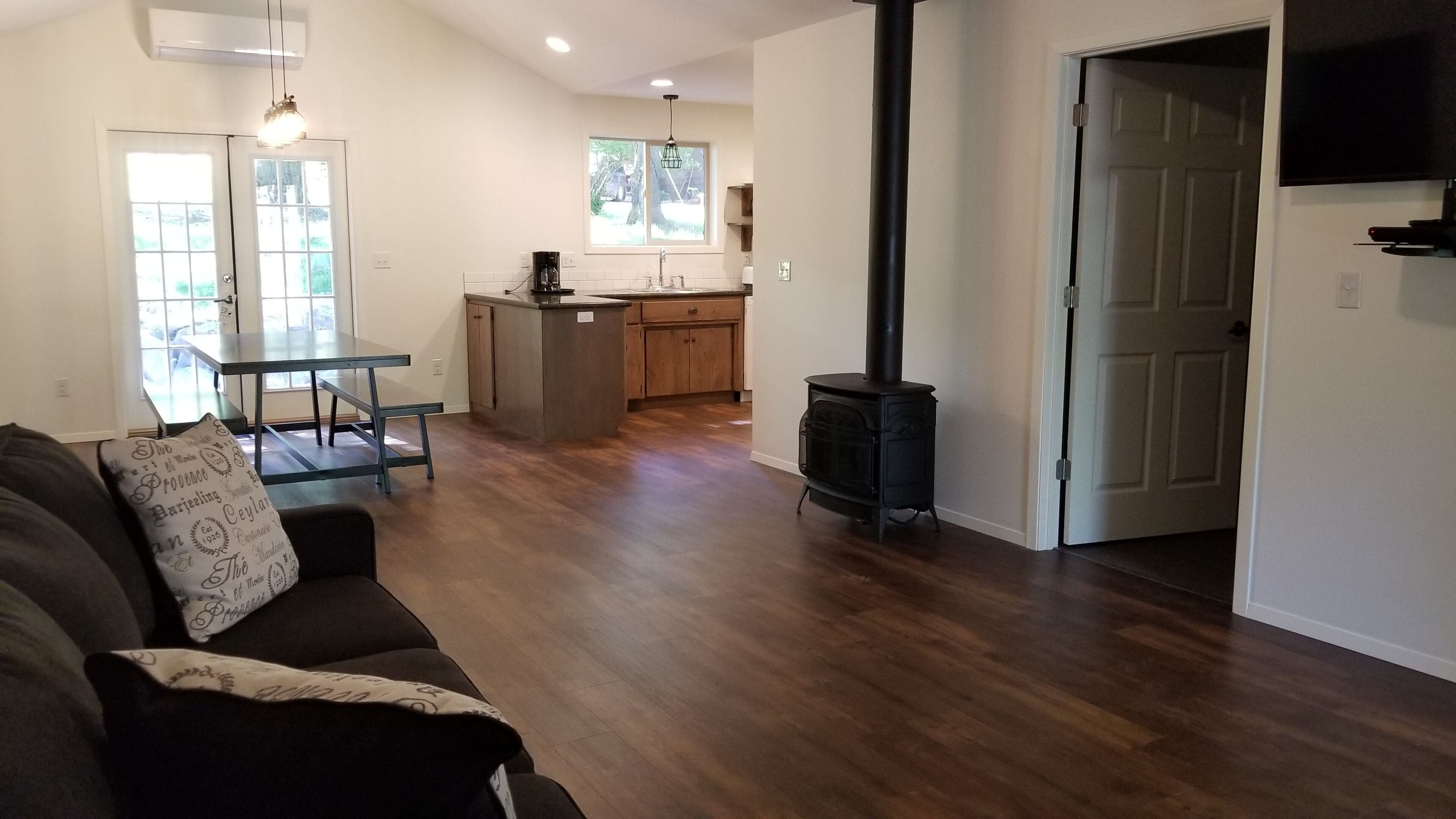 A living room with a sofa, a coffee table, and a fireplace. There is a kitchen area with a window and a door leading to another room.