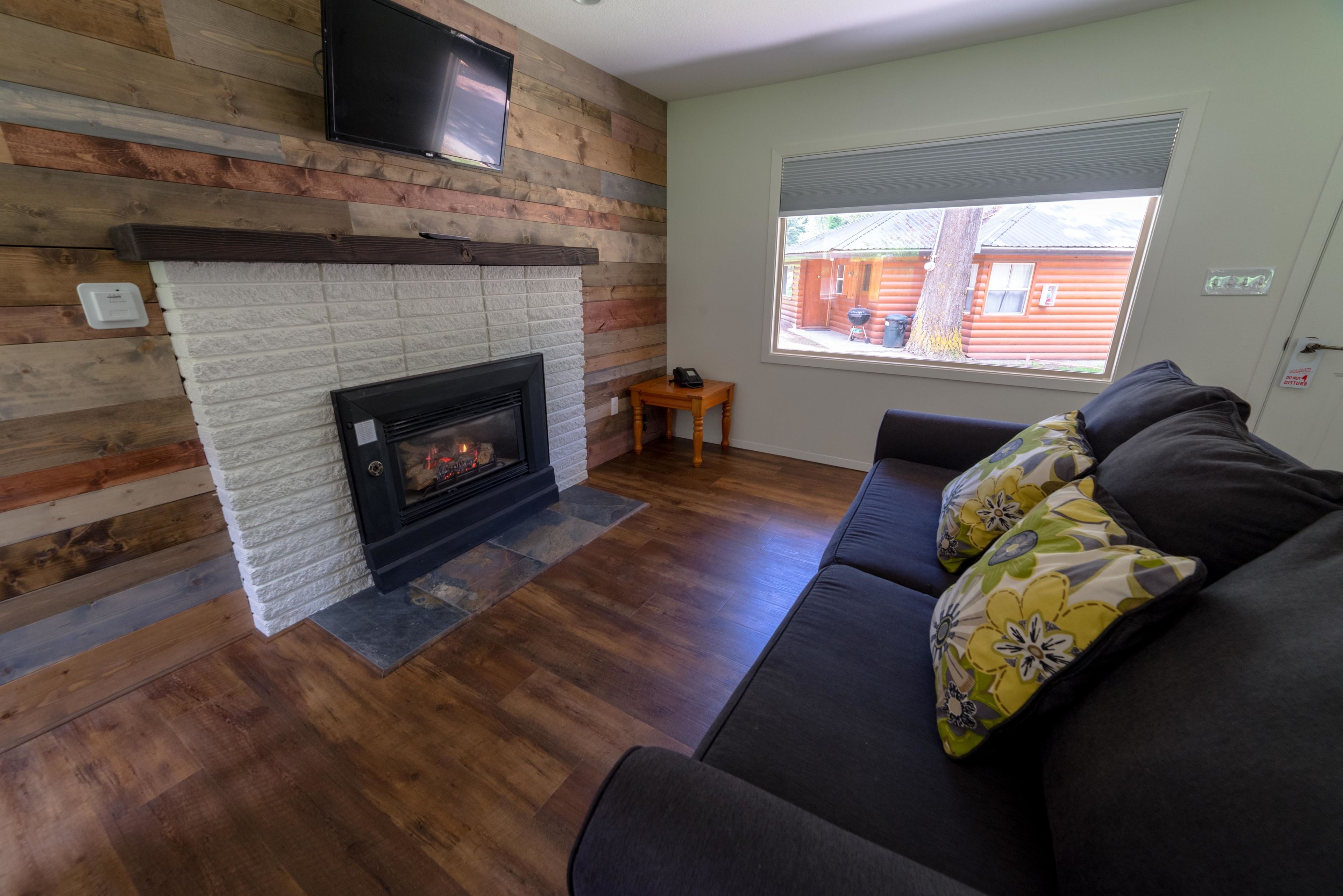 A living room with a fireplace, a sofa, and a window with a view of a cabin.