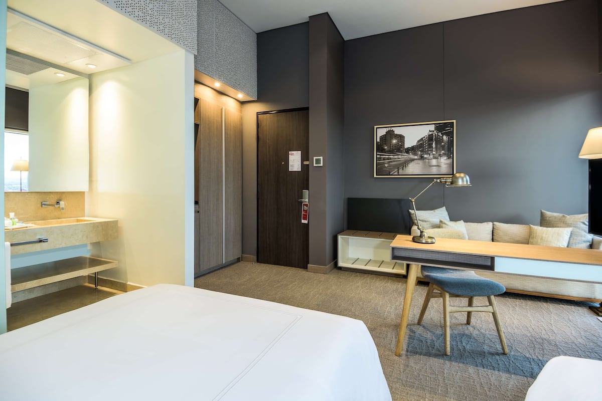 Habitación de hotel con cama, escritorio, silla, lámpara de escritorio, espejo, lavabo y un cuadro en la pared.