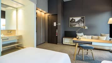 Quarto de hotel com cama, escrivaninha, cadeira, luminária, espelho, pia e quadro na parede.