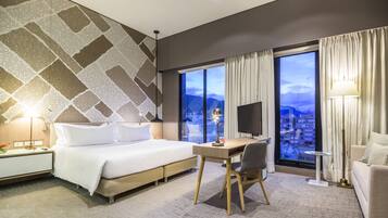 A hotel room with a large bed, a desk, a chair, a TV, and a view of the city through large windows.