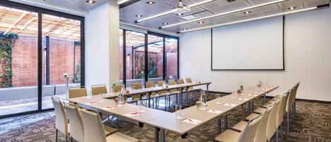 A conference room with a long table, chairs, a projector screen, and large windows with a view of a brick building.
