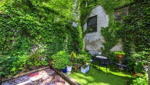 A lush garden with a brick pathway, a small seating area with a table and chairs, and a building partially covered in ivy. - Chelsea Pines Inn (New York)