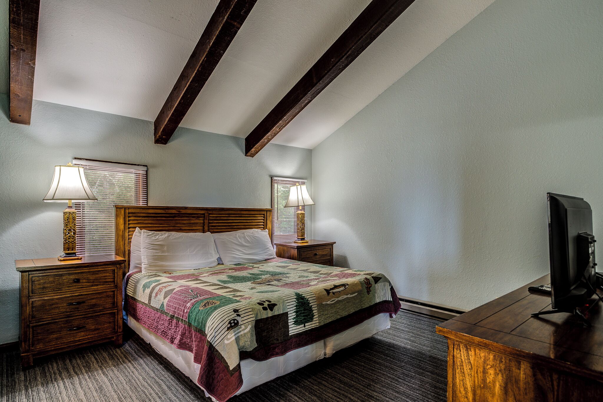 A neatly made bed with a quilt, two bedside lamps, a wooden nightstand, a desk with a computer, and a window with blinds.