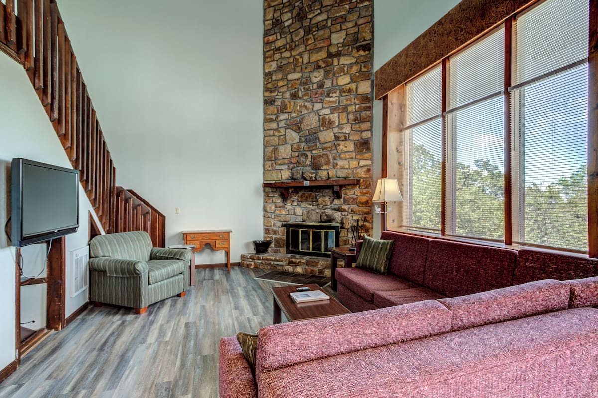 A living room with a stone fireplace, a television mounted on the wall, and a large window with blinds.