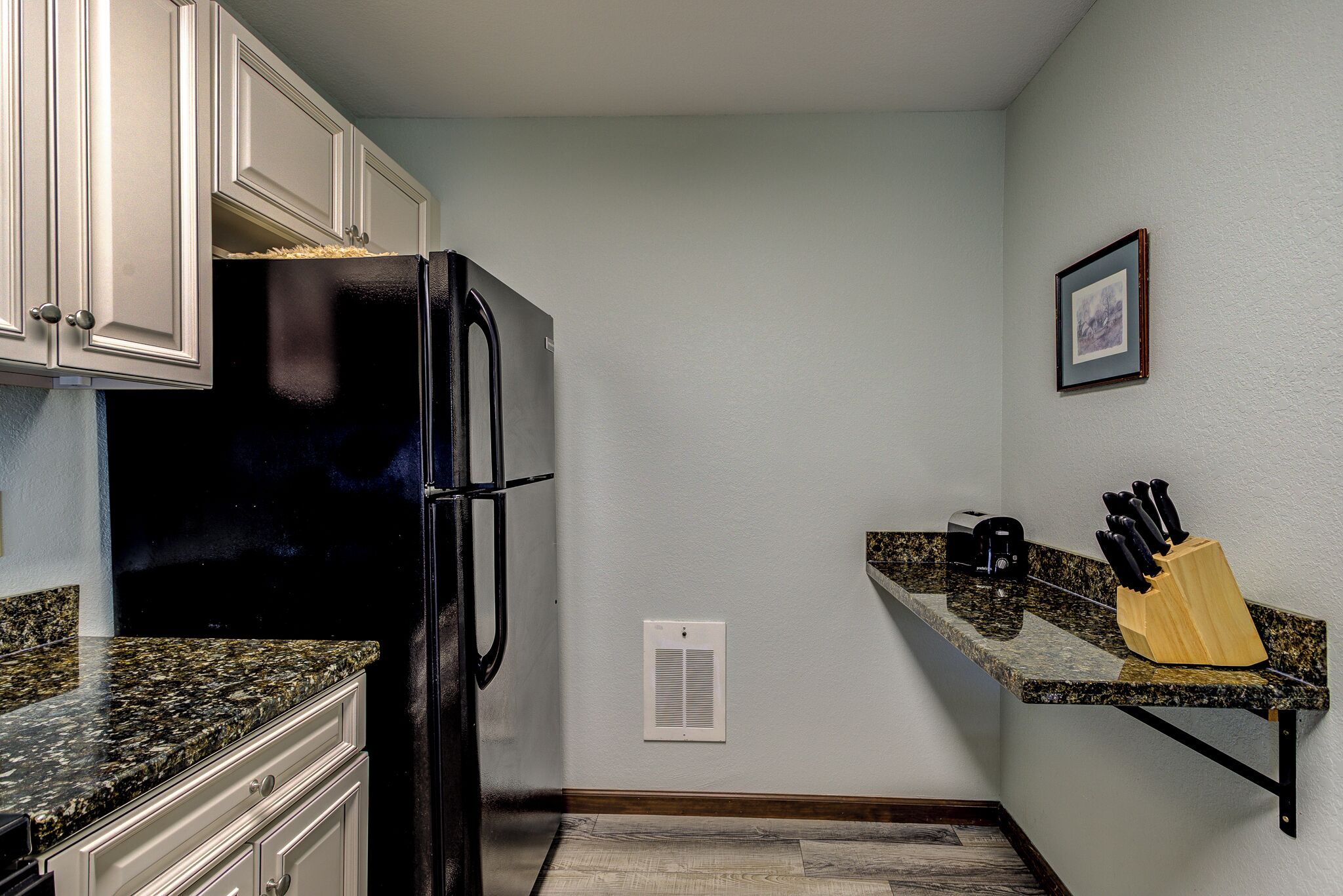 A kitchen with white cabinets, a black refrigerator, granite countertops, a wooden cutting board with knives, and a framed picture on the wall.