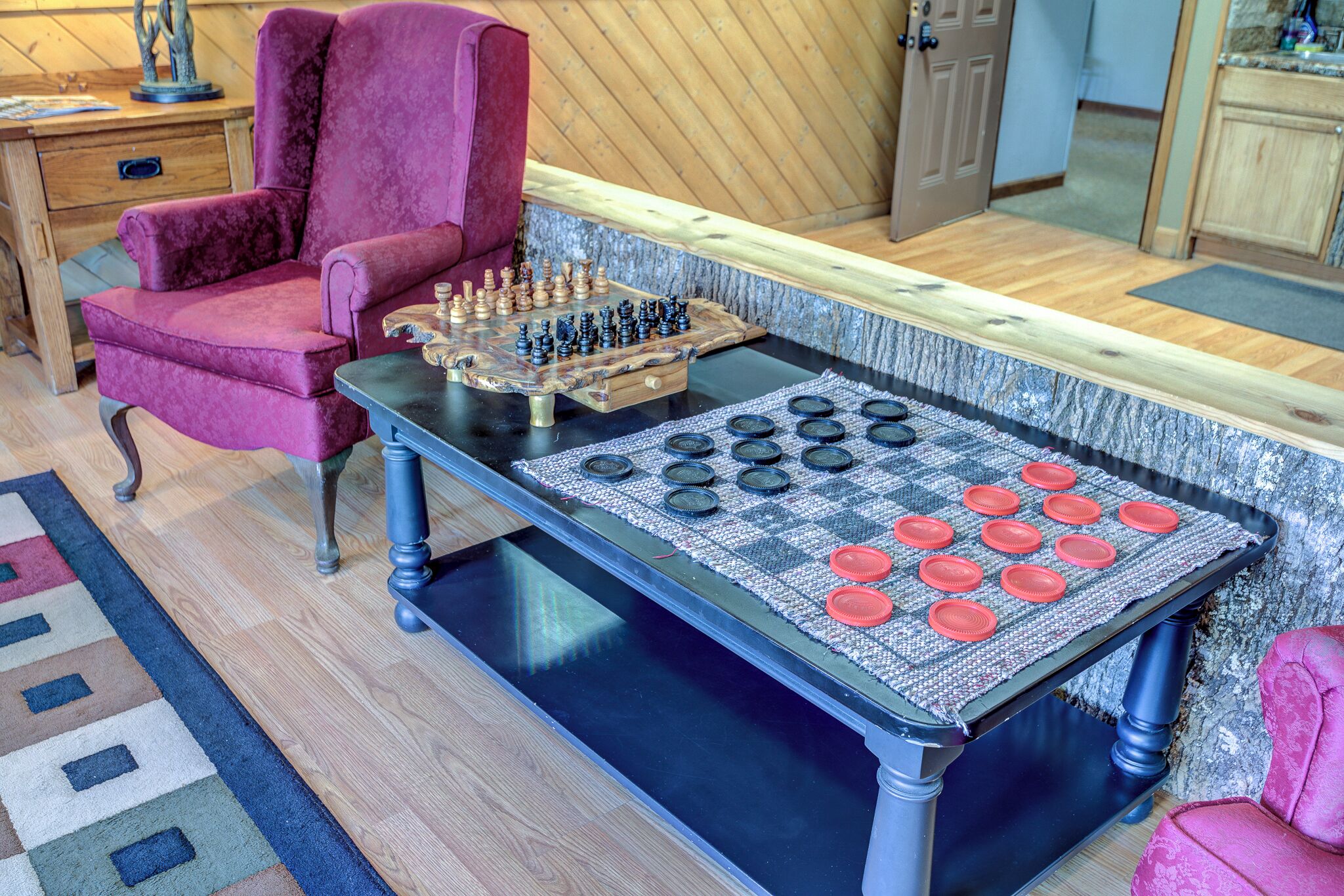 A checkered game board with pieces, a red armchair, and a wooden table in a room with wooden flooring and a door in the background.