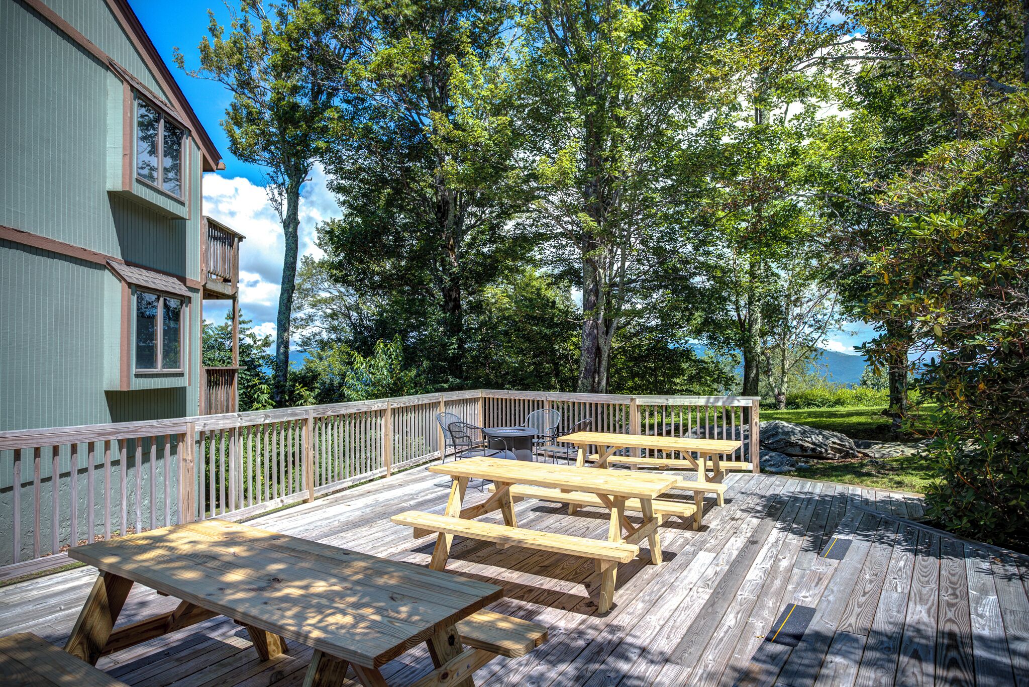 A wooden deck with picnic tables and a railing, surrounded by trees and a building.