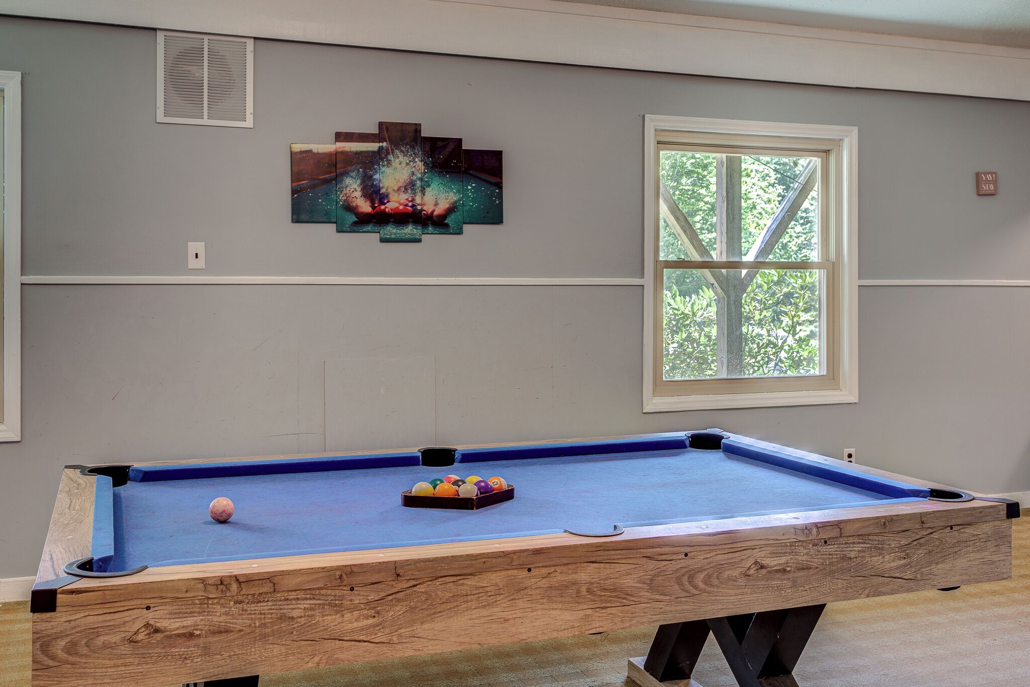 Uma mesa de bilhar com bolas, uma janela com vista para árvores e uma obra de arte pendurada na parede.