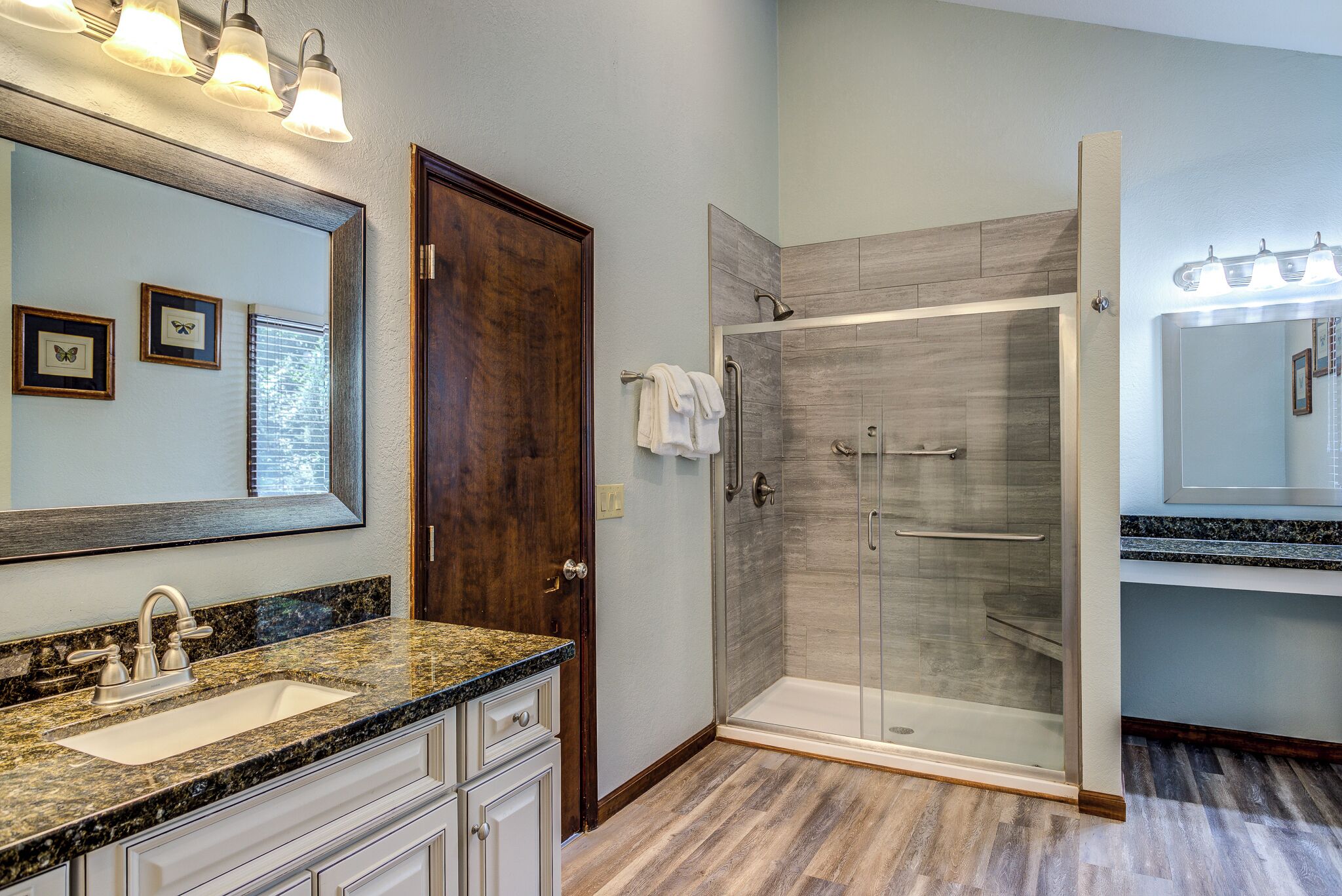 A bathroom with a granite countertop sink, a large mirror, a wooden door, and a glass shower enclosure.