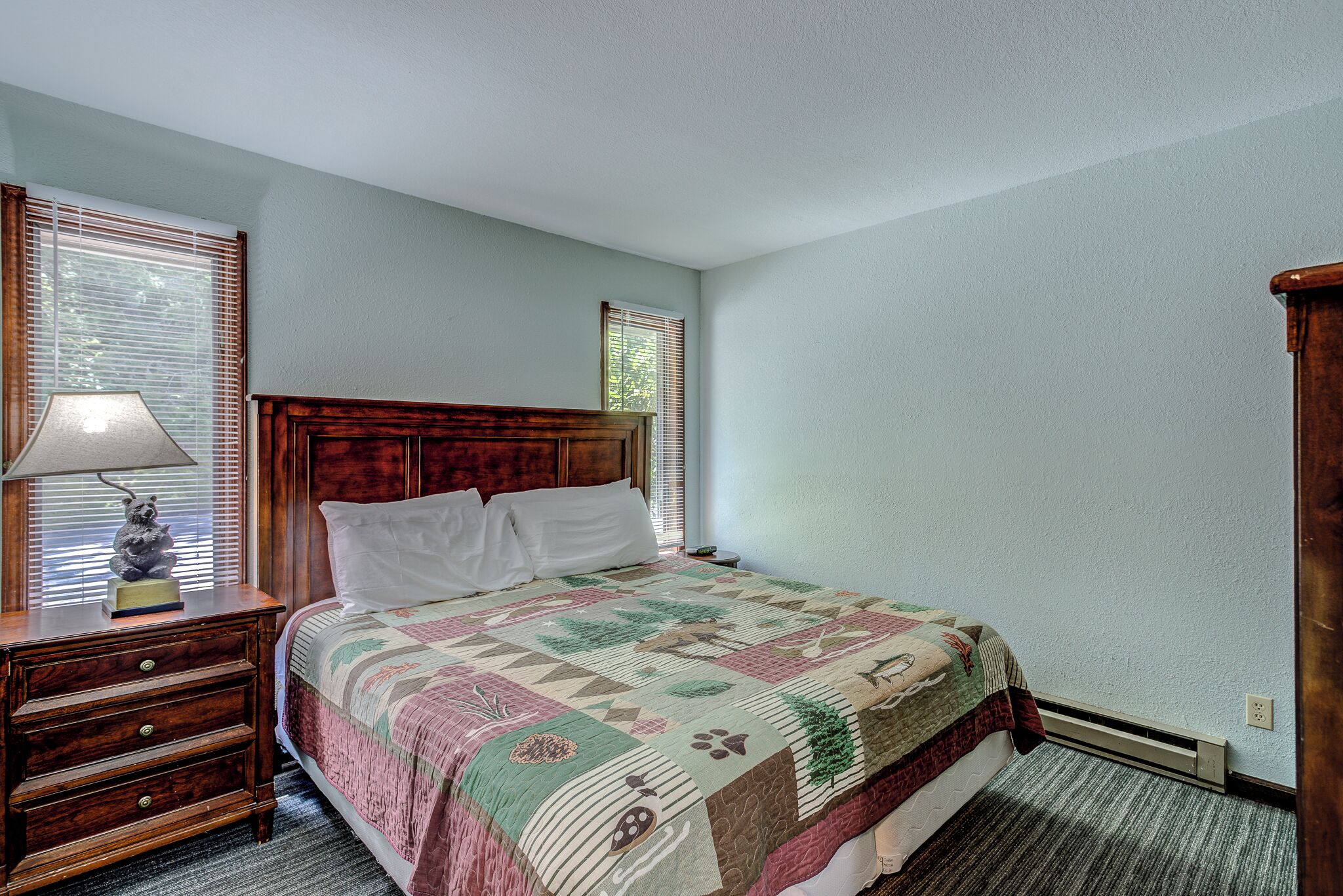 A bedroom with a wooden headboard, a quilted bedspread, a nightstand with a lamp, and a dresser.