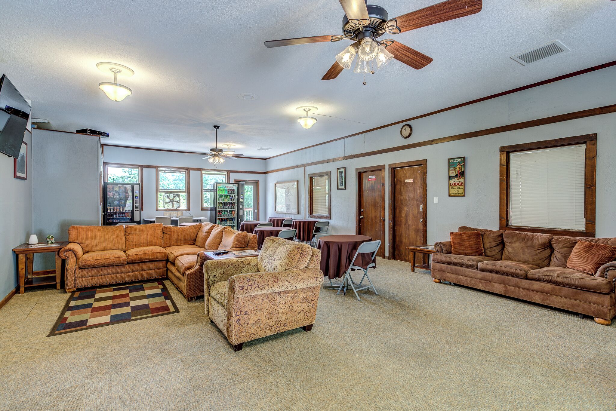 A spacious living room with a large sofa, a smaller armchair, a coffee table, and a dining table with chairs. There is a ceiling fan, recessed lighting, and a clock on the wall.