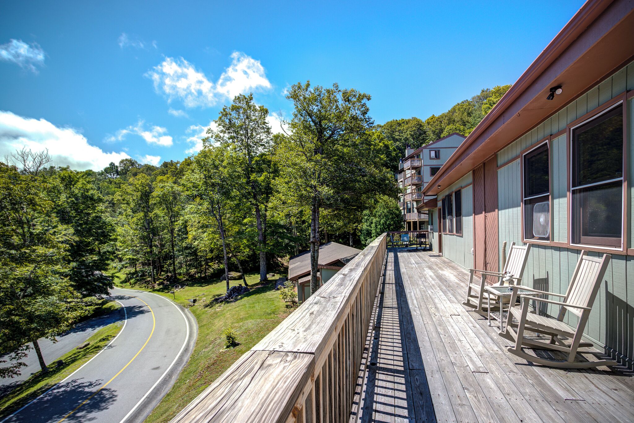 A wooden deck with rocking chairs overlooking a curved road and dense forest.