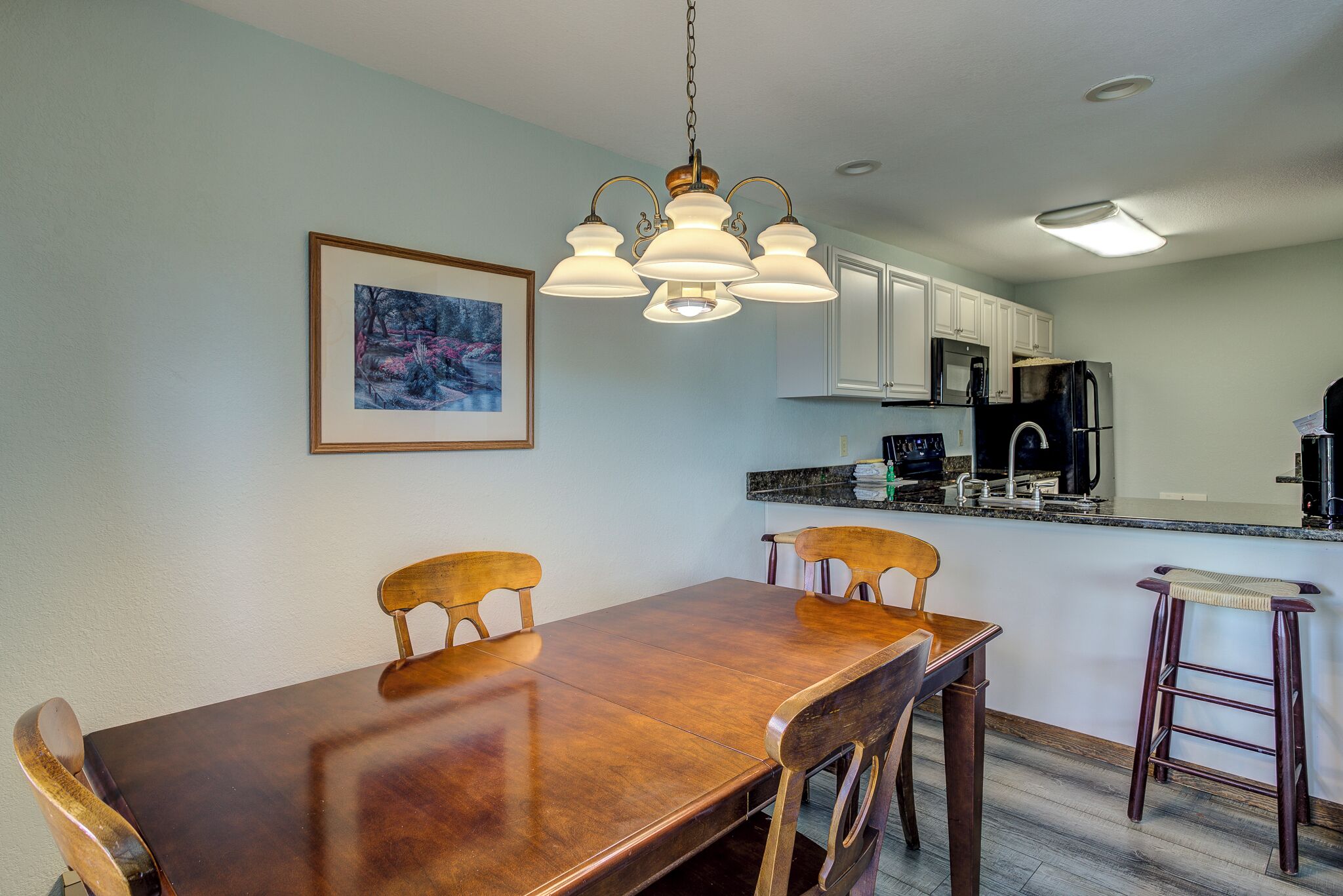 A dining area with a wooden table and chairs, a hanging light fixture, and a kitchen in the background.