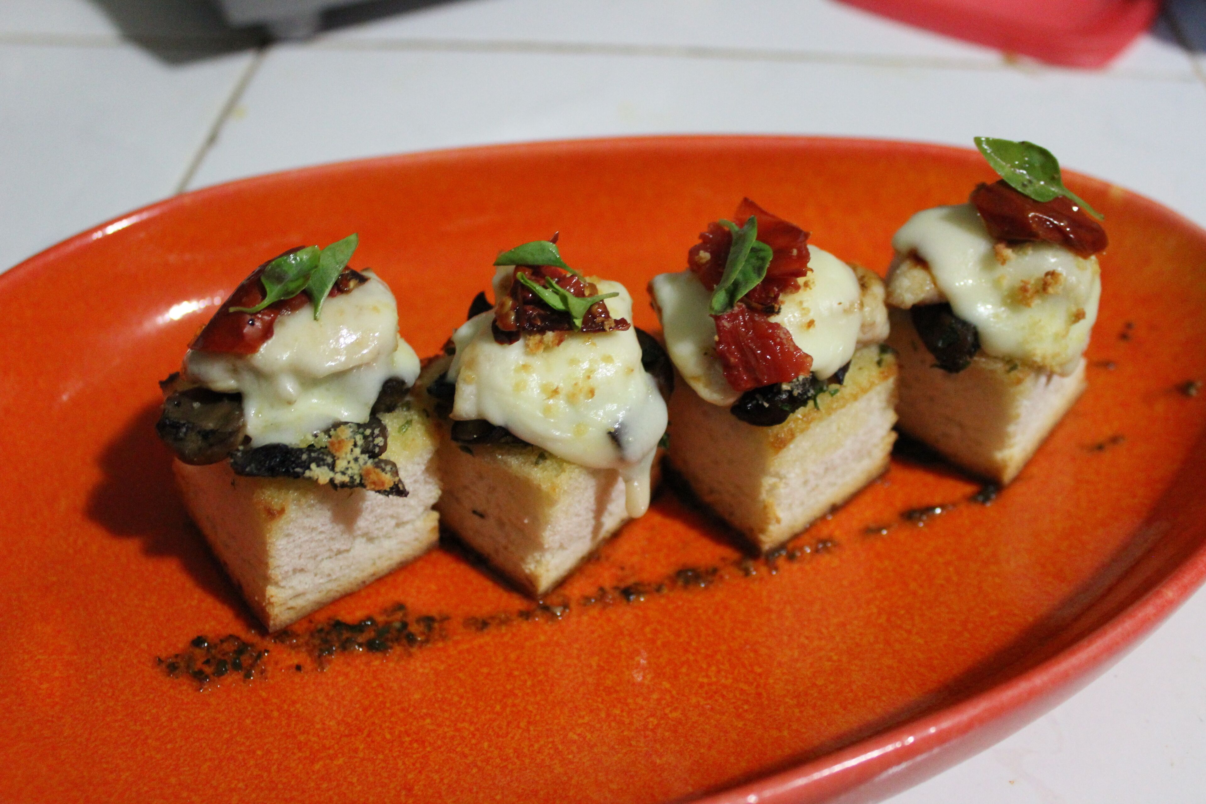 A red plate with four pieces of food topped with cheese, herbs, and possibly bacon.