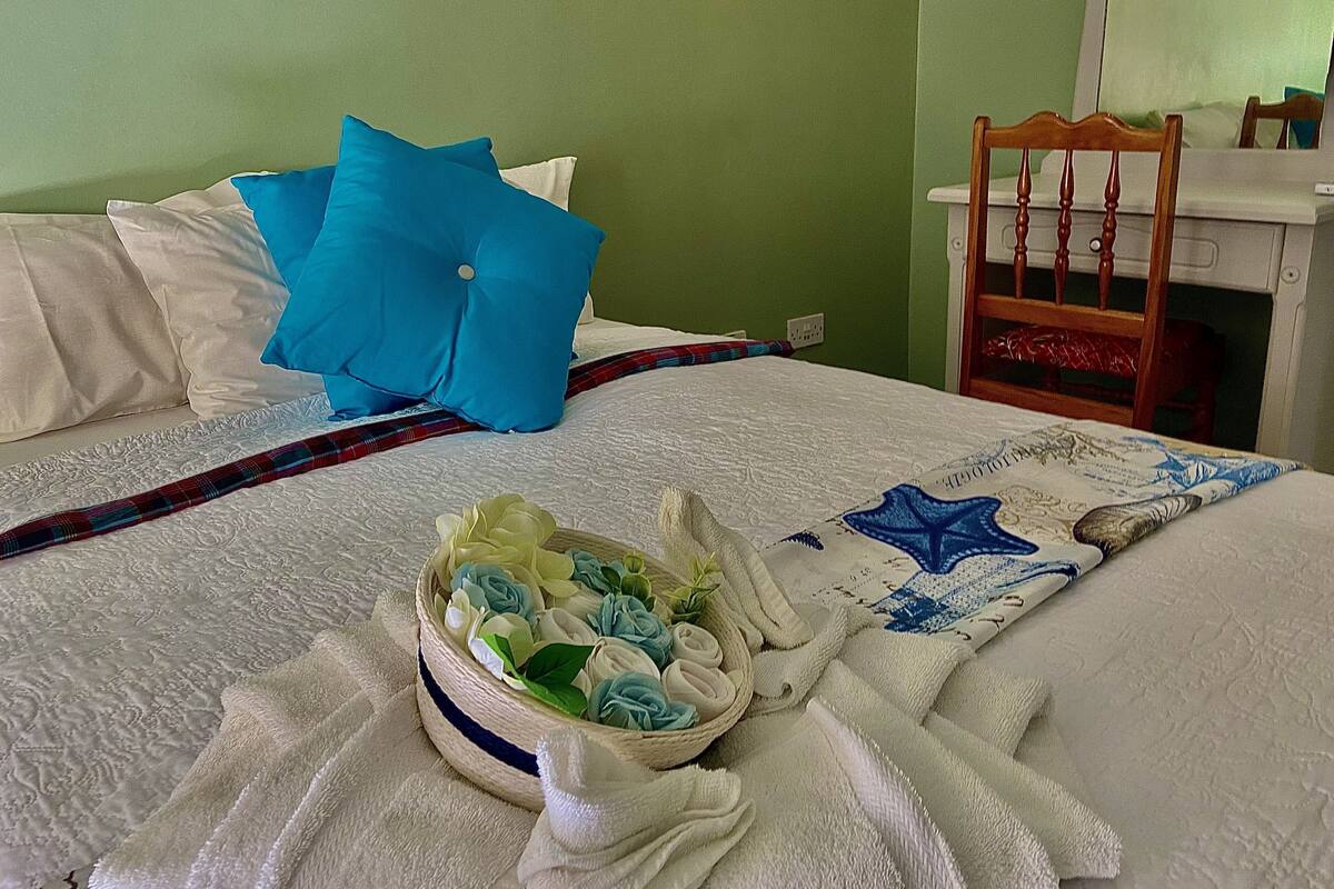 A neatly made bed with a blue pillow, a white pillow, and a plaid blanket. There is a basket of folded towels and a starfish-patterned blanket on the bed.