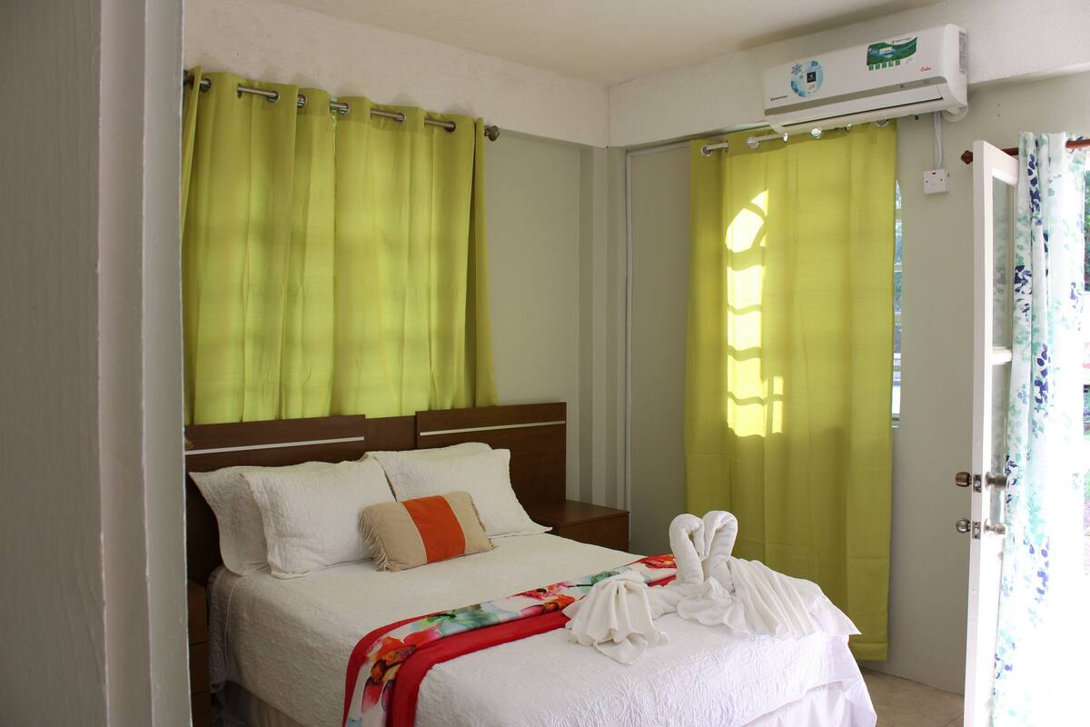 A hotel room with a bed, a nightstand, a window with curtains, and an air conditioning unit.