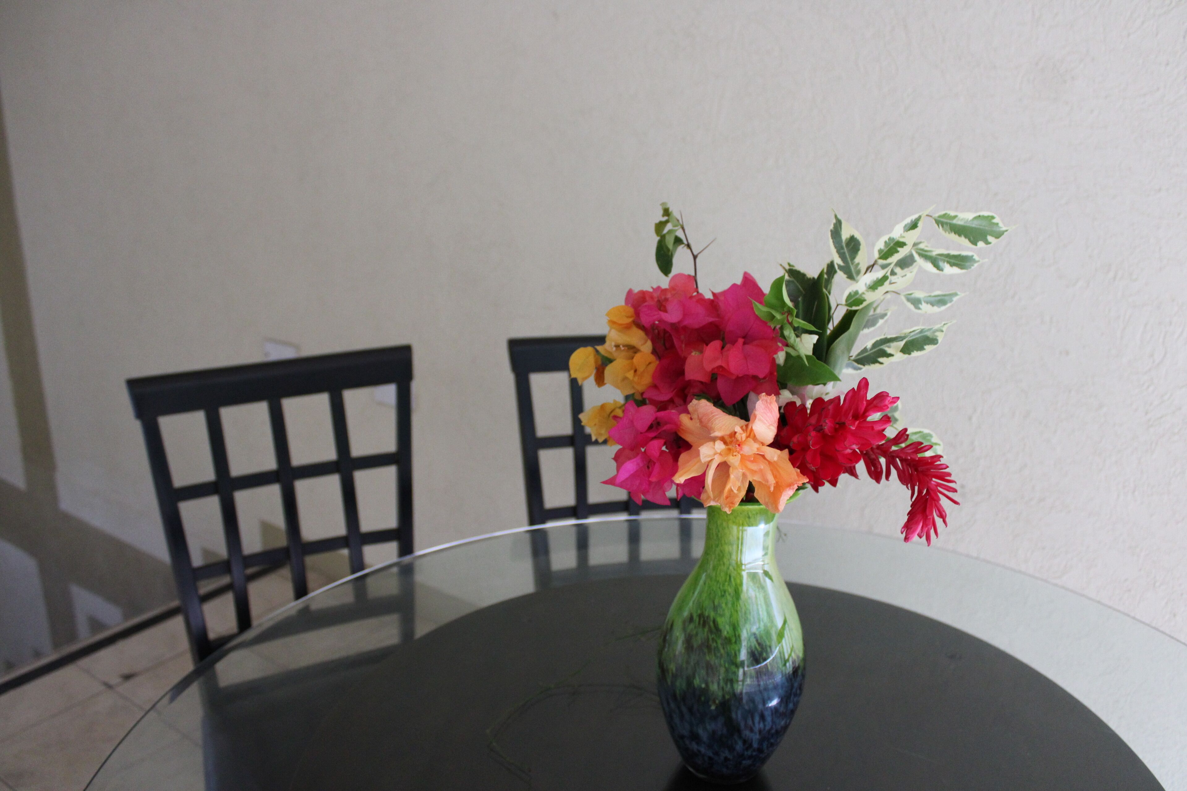 Une table ronde en verre sur laquelle est posé un vase vert contenant des fleurs aux couleurs vives.