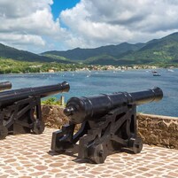 Dos cañones sobre una plataforma de piedra con vista a una bahía con barcos y montañas al fondo.