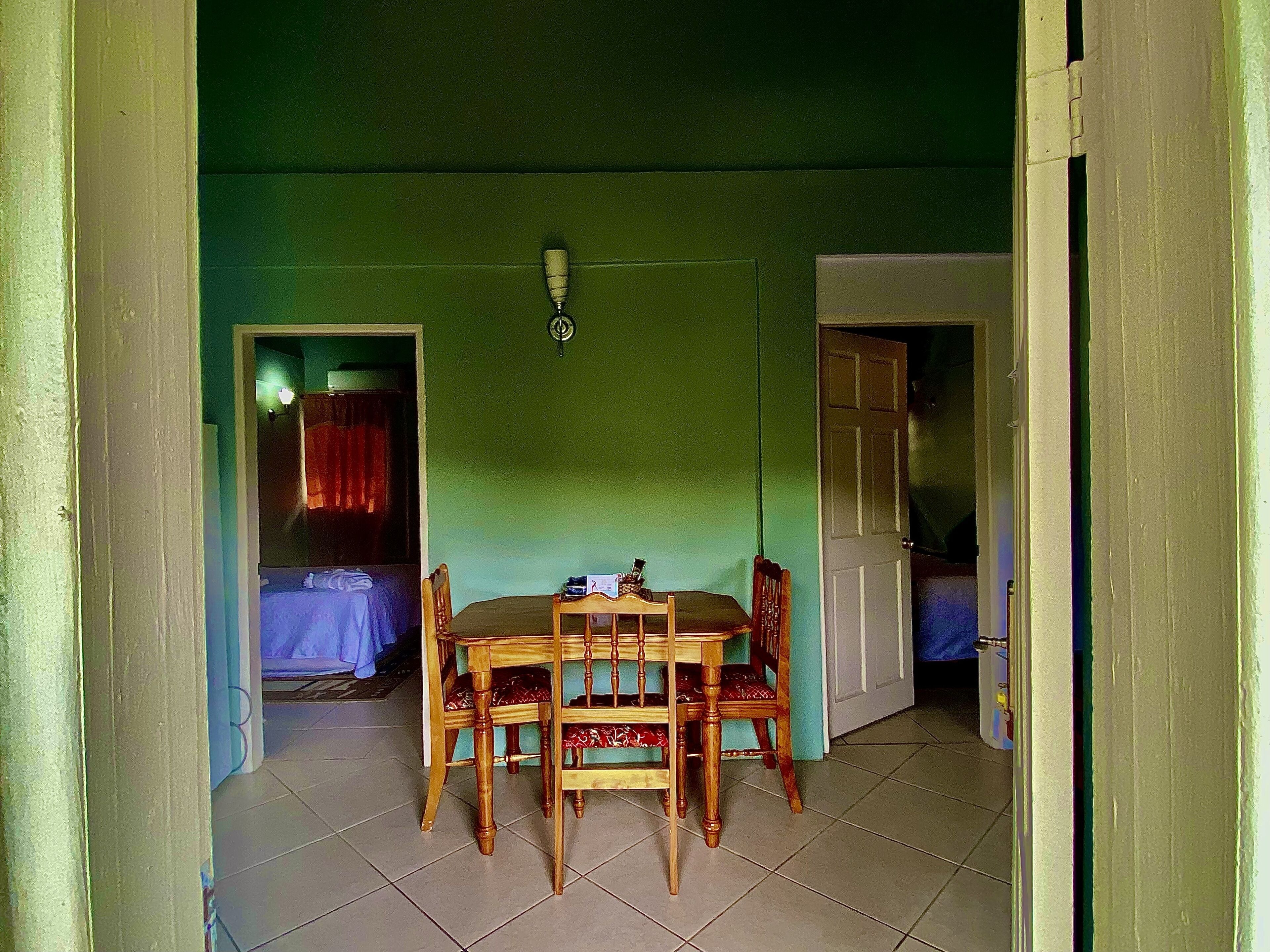 Uma área de refeições com mesa e cadeiras de madeira, luminária no teto e vista para outro quarto com cama e cortinas.