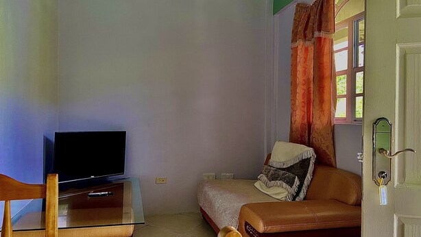 A small, sparsely furnished room with a television on a glass table, a single bed with a patterned blanket, and a window with curtains. - Rejens Hotel (Portsmouth)