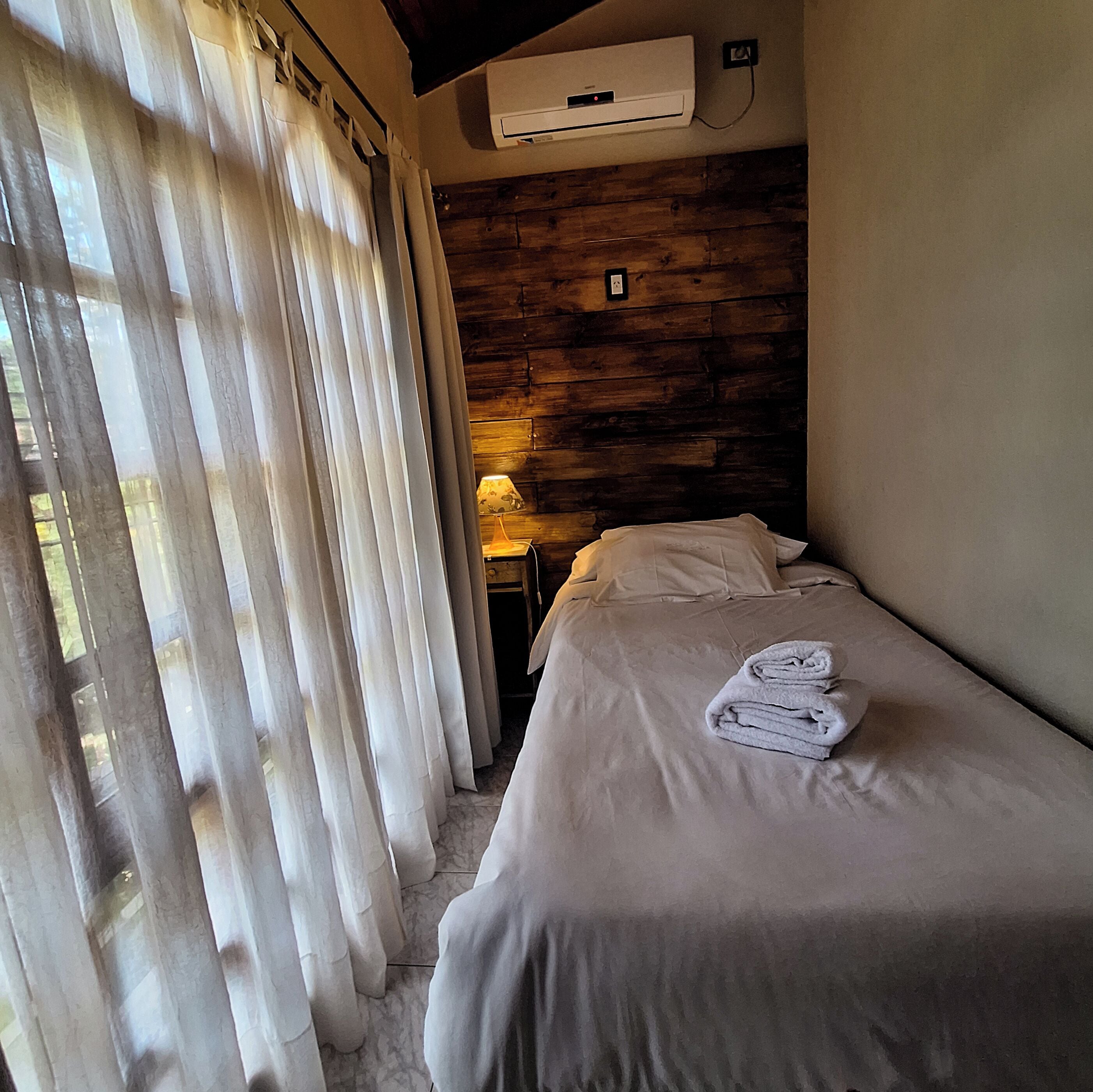 Habitación de hotel con cabecera de madera, cama con ropa blanca y ventana con cortinas traslúcidas.