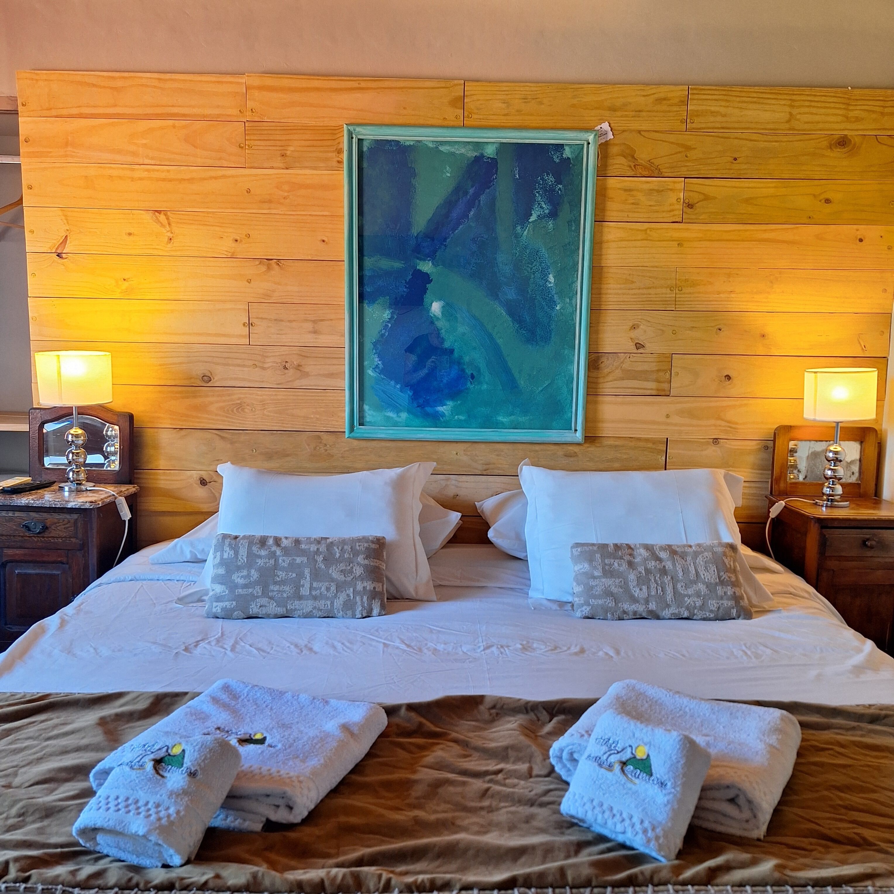 A hotel room with a wooden headboard, a large abstract painting, two bedside lamps, a bed with white linens, and two towels on the bed.