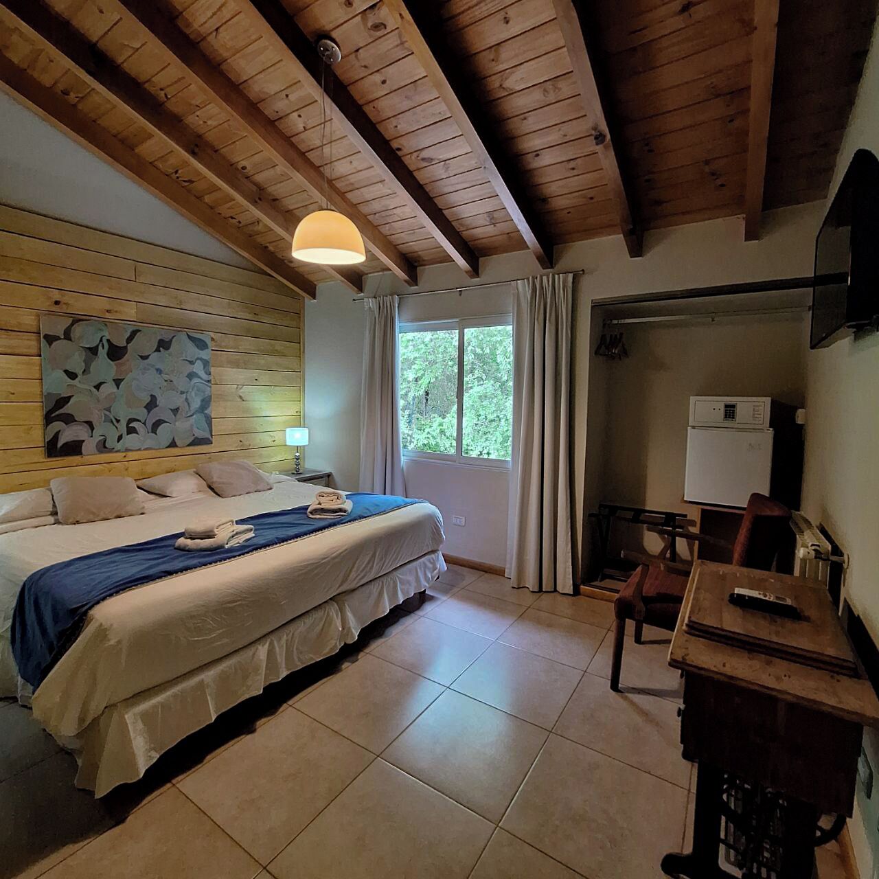 A room with a bed, a desk, a chair, a window with curtains, a ceiling fan, and a wall-mounted TV.