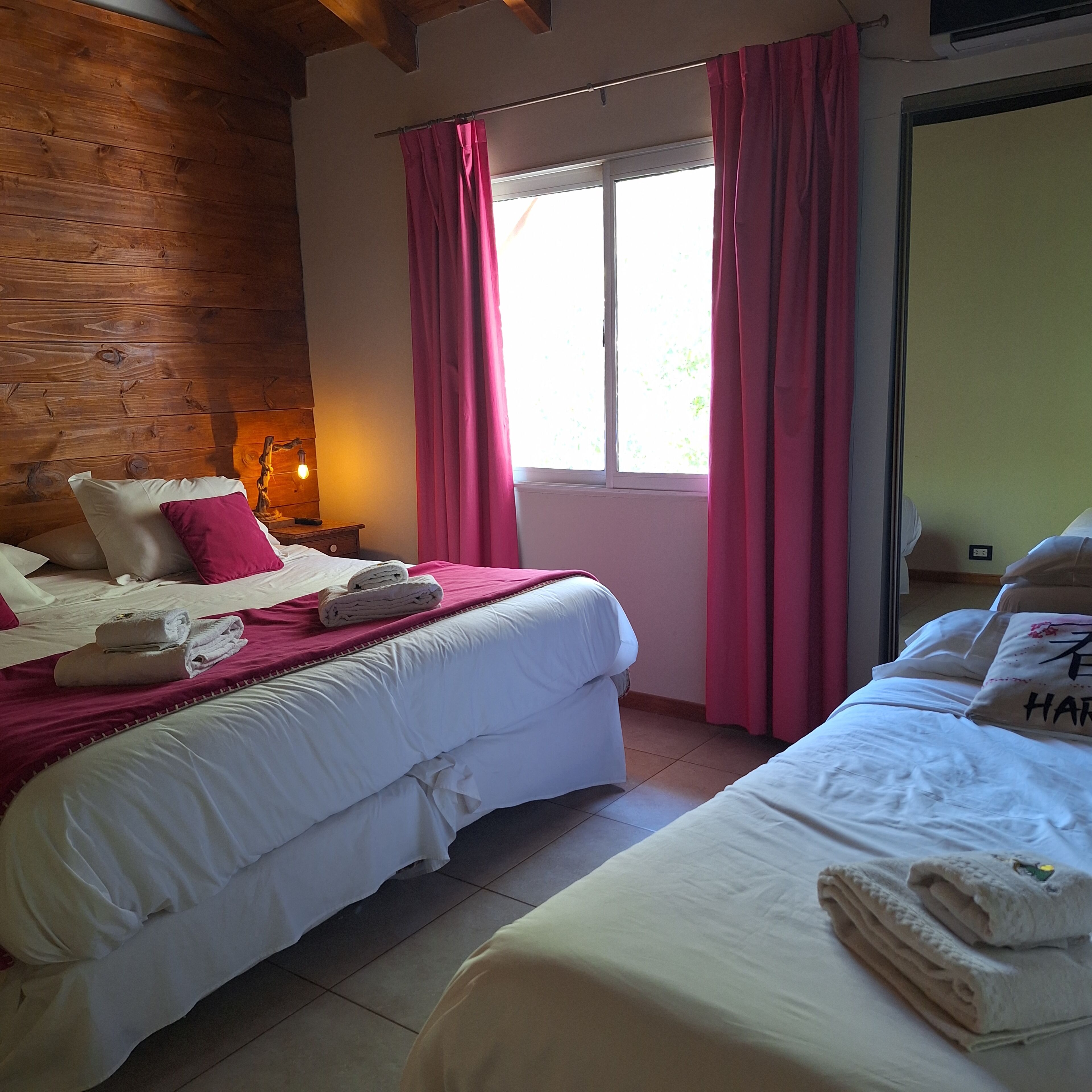 Habitación de hotel con dos camas, paredes de madera, una ventana con cortinas rosadas y una mesita de noche con lámpara.