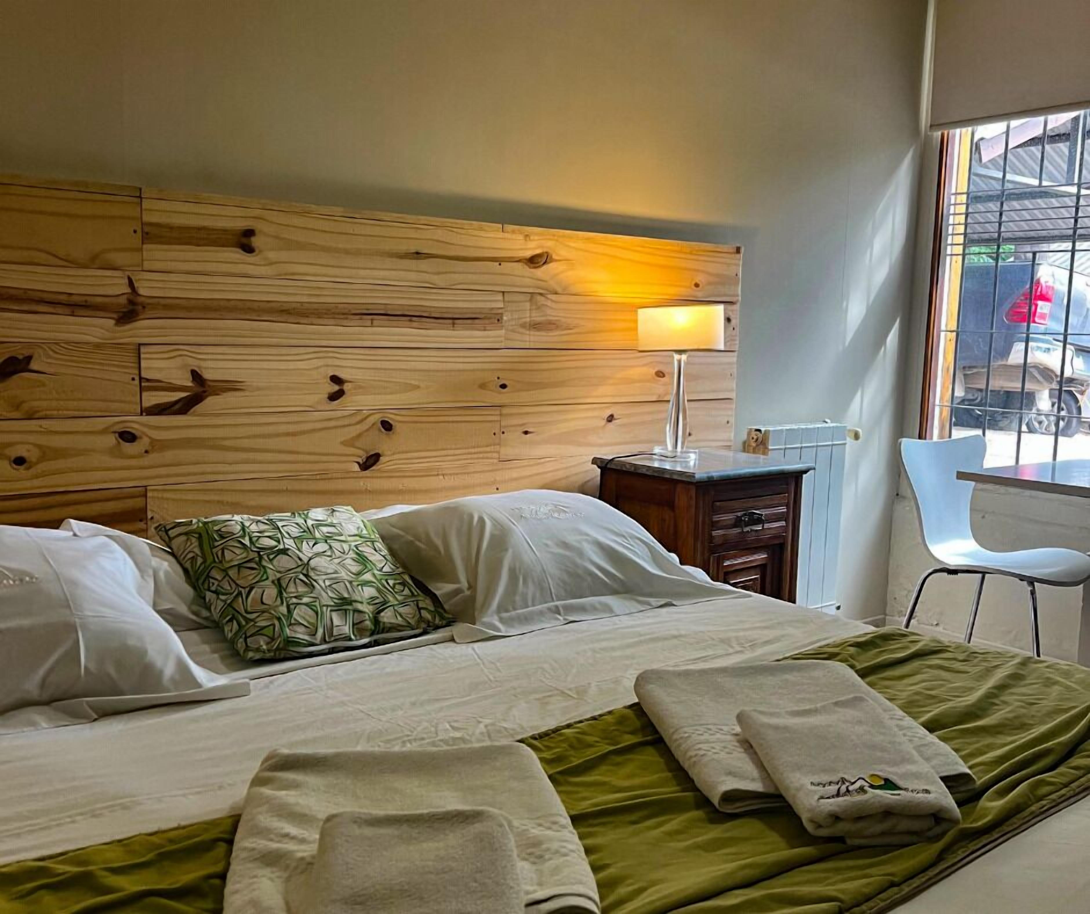 A hotel room with a wooden headboard, a bedside table with a lamp, a green bedspread, and a view of the outside.