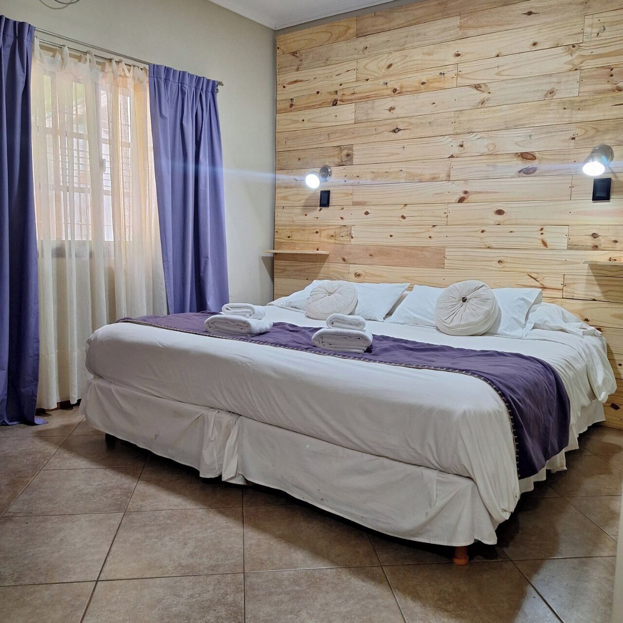 Ein Schlafzimmer mit einem hölzernen Kopfteil, einem Bett mit weißer Bettwäsche, einer violetten Decke und einem Fenster mit blauen Vorhängen.