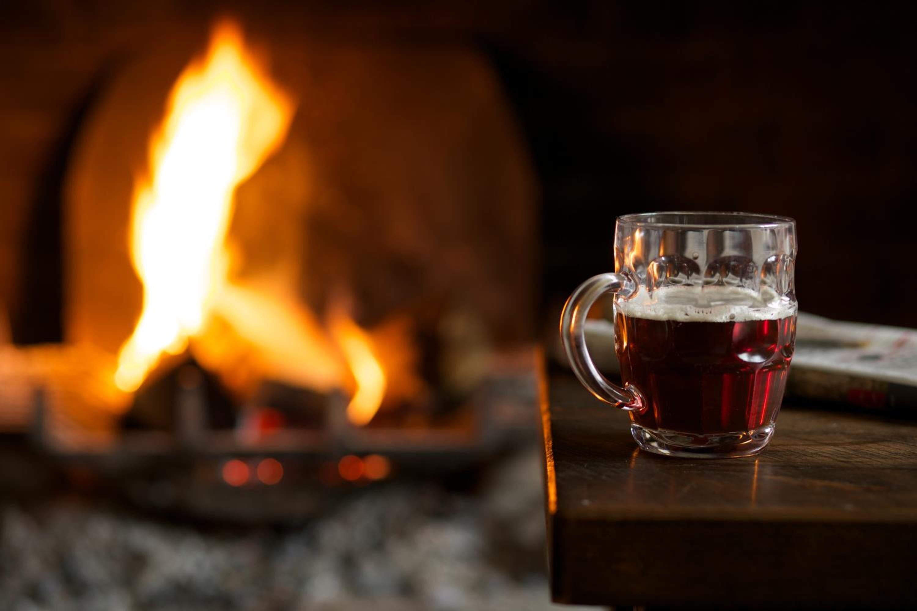 Una taza de vidrio con una bebida sobre una superficie de madera, con una chimenea desenfocada al fondo.