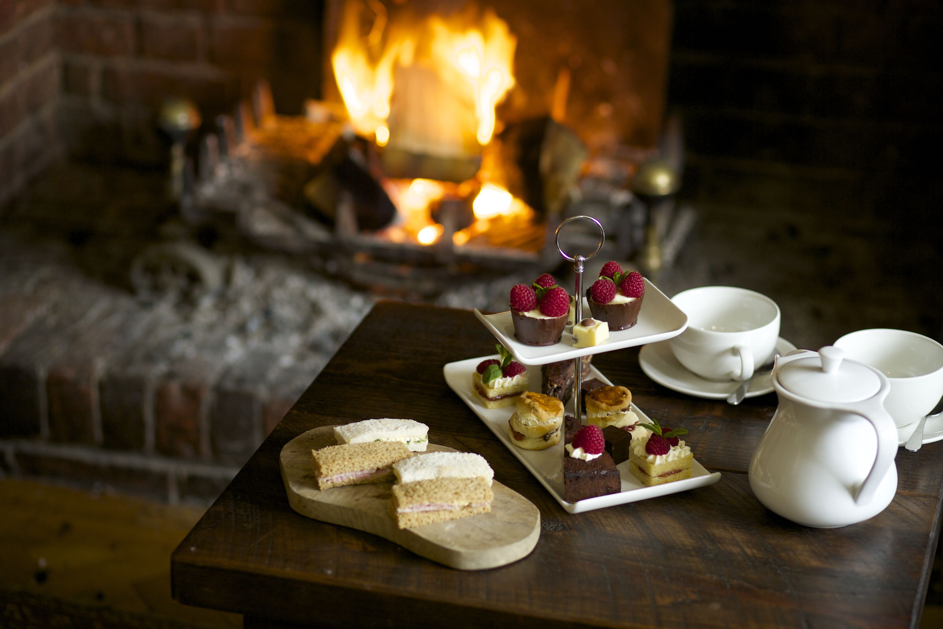 A table with various food items and white cups and a teapot, set against a brick fireplace with a visible fire.