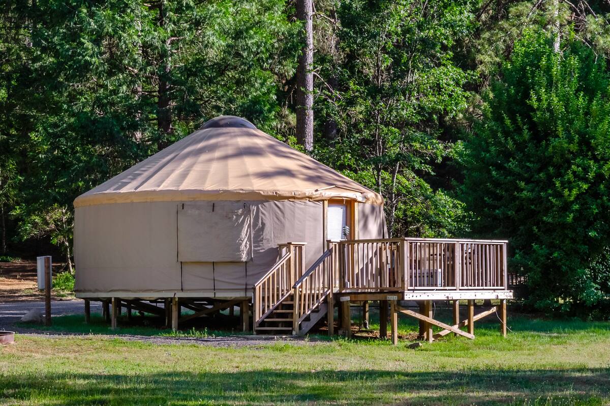 A single-story, A-frame yurt with a wooden deck and stairs, surrounded by dense forest.