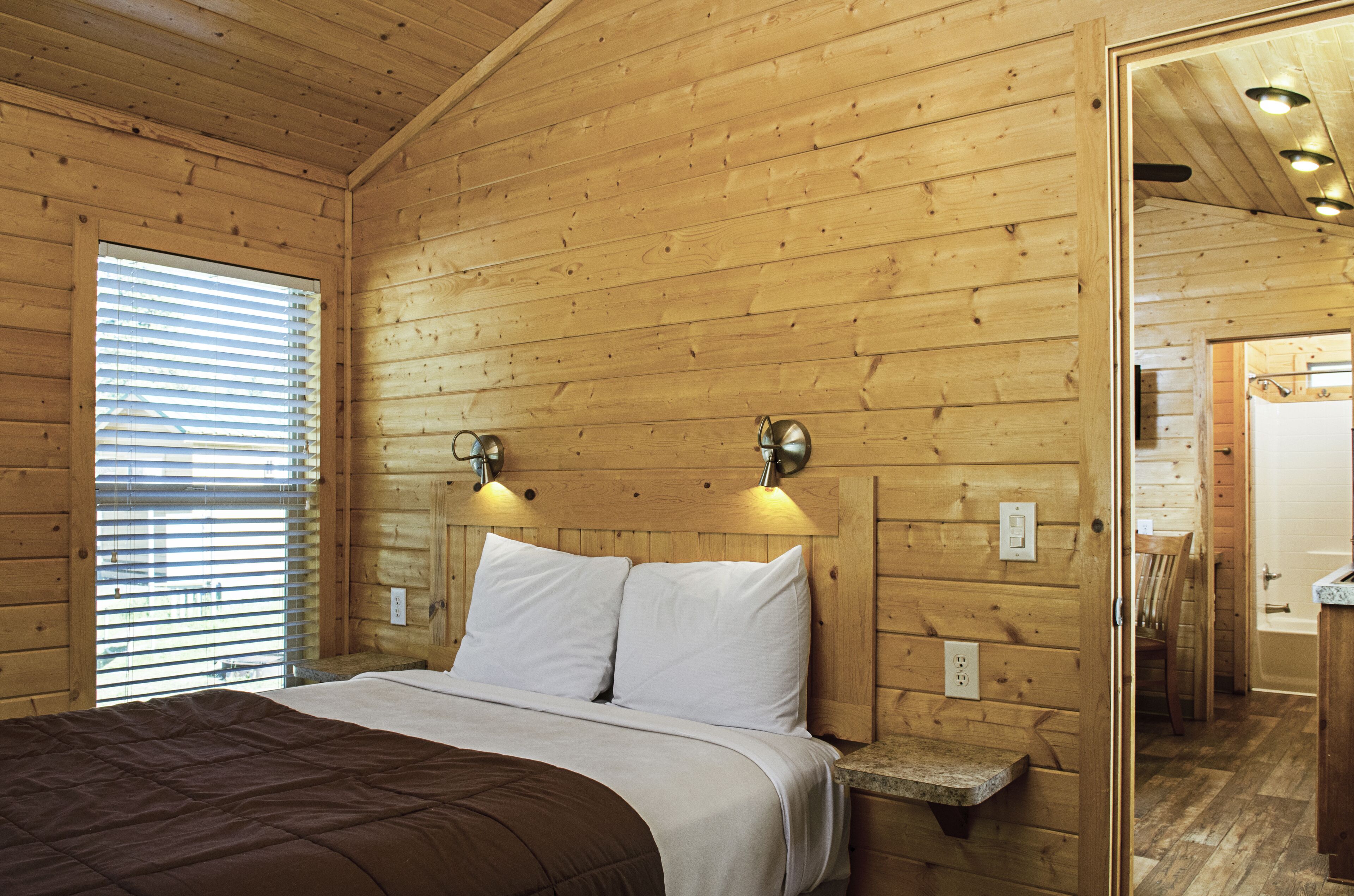 Habitación de cabaña de madera con una cama, dos luces fijadas a la pared, un ventanal con persianas y una puerta de baño visible.