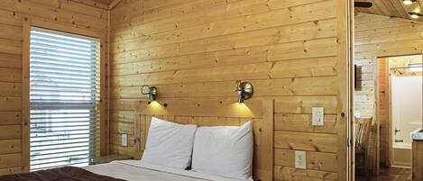Habitación de cabaña de madera con una cama, dos luces fijadas a la pared, un ventanal con persianas y una puerta de baño visible.