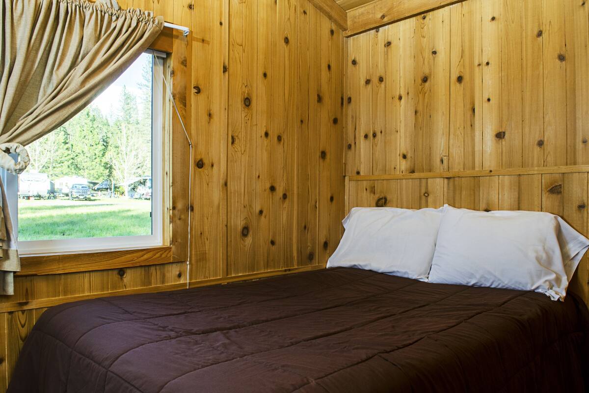 A wooden room with a bed, a window with curtains, and a view of a grassy area outside.