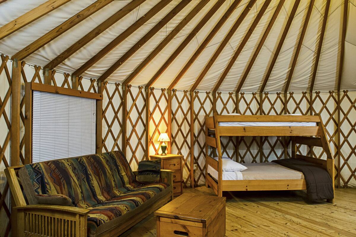 A cozy interior of a tent with a sofa, a wooden bed, a small table, and a window with blinds.